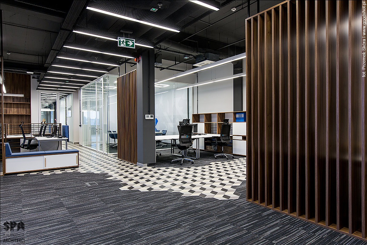 Nowoczesne biuro z wykładziną dywanową w odcieniach szarości, przechodzącą w geometryczny wzór płytek na podłodze, drewniane lamele działowe, szklane ścianki oddzielające przestrzenie biurowe.