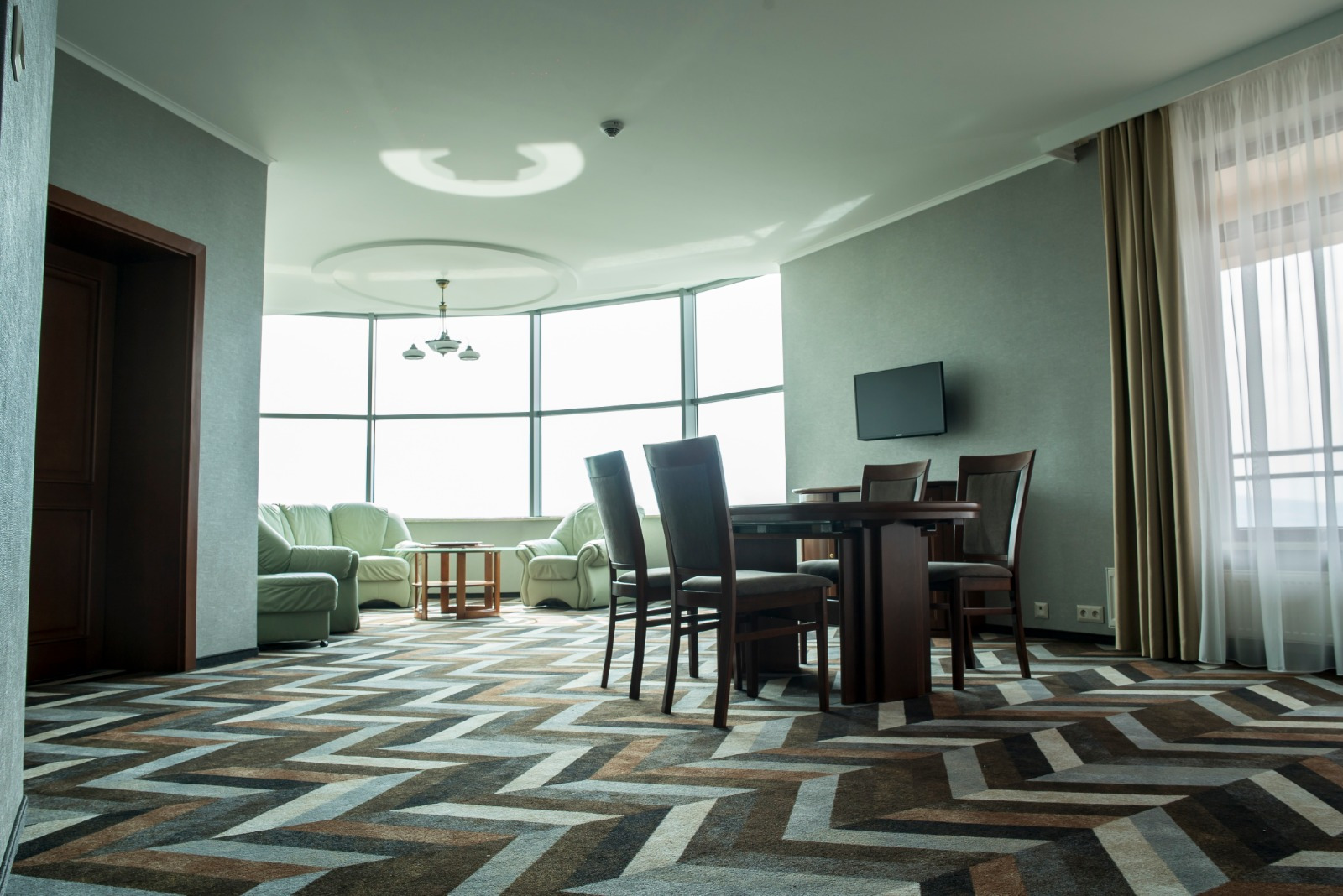 Wnętrze pokoju z wzorzystą podłogą w zygzaki, ciemnobrązowy stół i krzesła, skórzane sofy w tle, panoramiczne okno z widokiem.