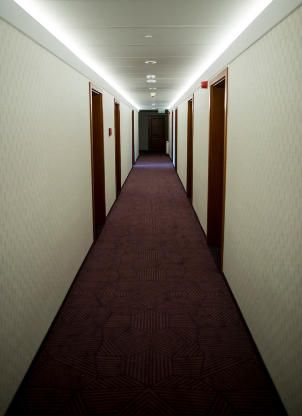 Długi hotelowy korytarz z wzorzystą, bordową podłogą i rzędem drzwi po obu stronach, oświetlony listwami LED w suficie.