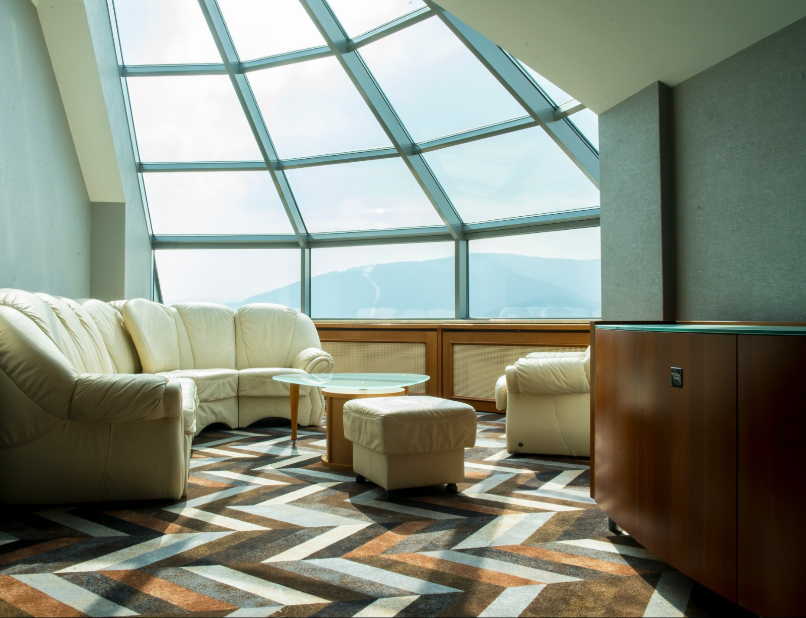 Jasny salon z wykładziną w zygzaki, skórzana sofa i panoramiczne okno z widokiem na góry.