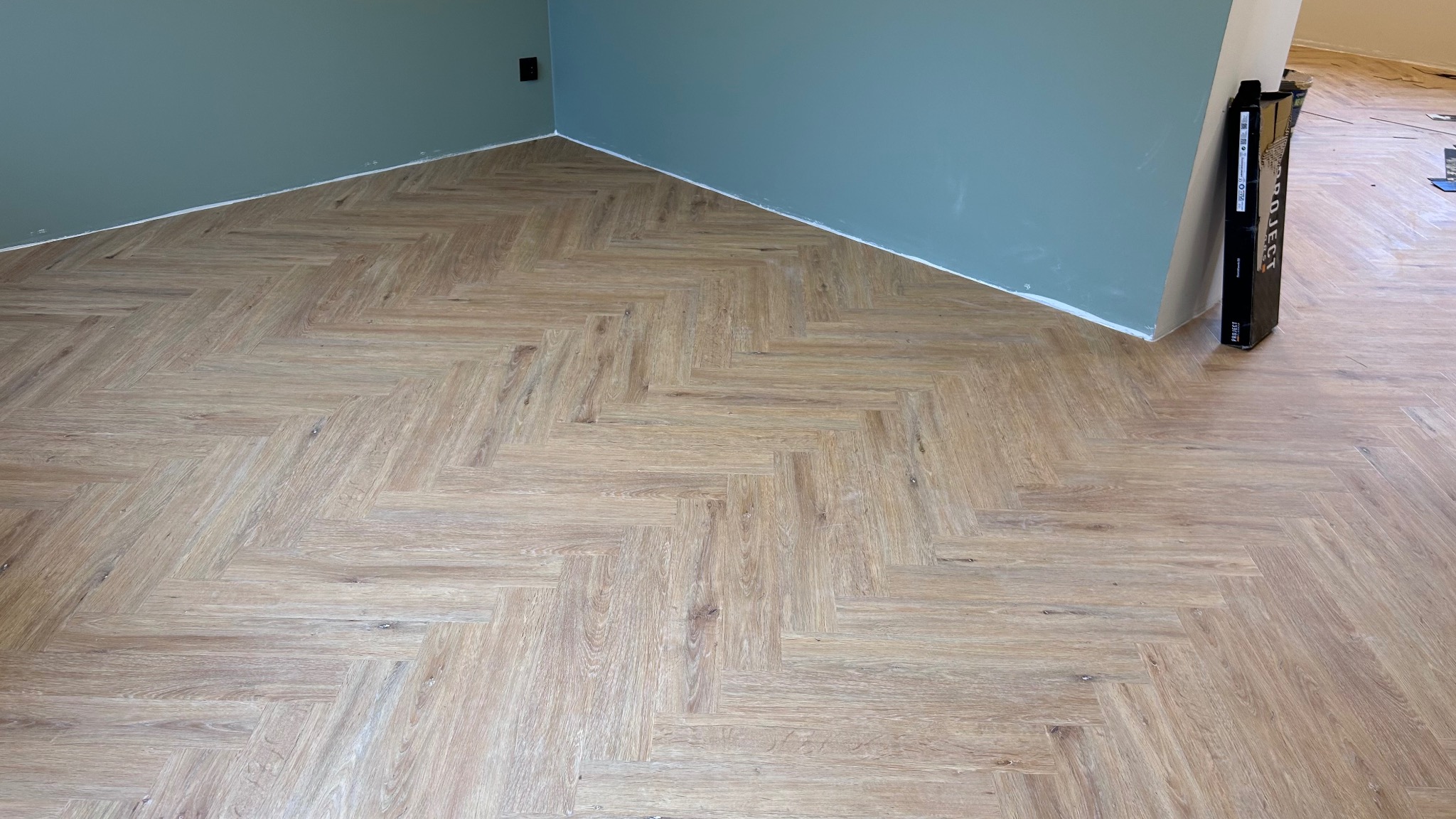 Ułożona podłoga w jodełkę z paneli o wzorze drewna, przy ścianie stoi karton z panelami