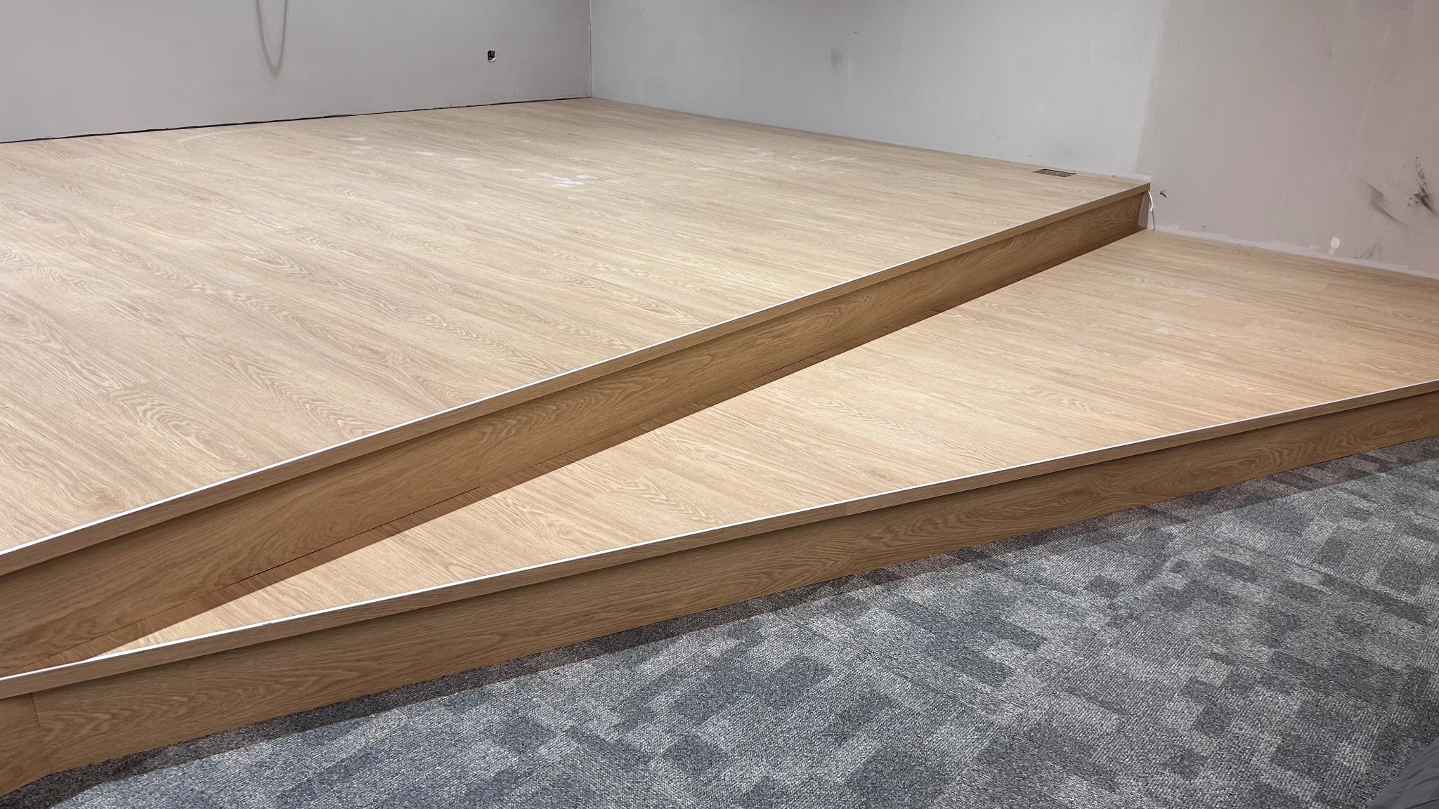 Dwu-poziomowa podłoga z panelami imitującymi jasne drewno, wykończona białą listwą przypodłogową, widoczna szara wykładzina dywanowa w dolnej części kadru, surowe ściany w tle.