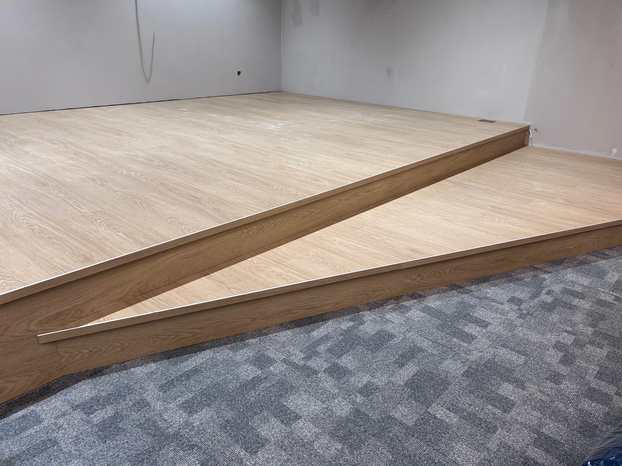 Podwyższona drewniana podłoga z dwoma stopniami, wykończona listwami przypodłogowymi, na tle szarej wykładziny dywanowej w geometryczny wzór, w surowym wnętrzu.