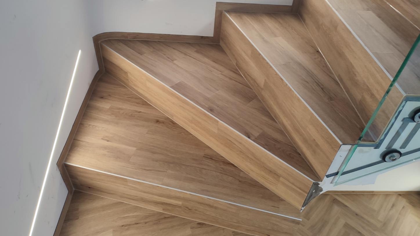 Schody wykończone panelami imitującymi drewno, z delikatnym oświetleniem LED przy ścianie i szklaną balustradą na górze, widok z góry.