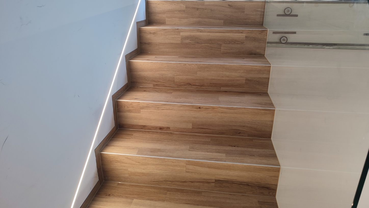 Schody wyłożone panelami imitującymi drewno, z subtelnym oświetleniem LED wzdłuż ściany, zakończone szklaną balustradą.