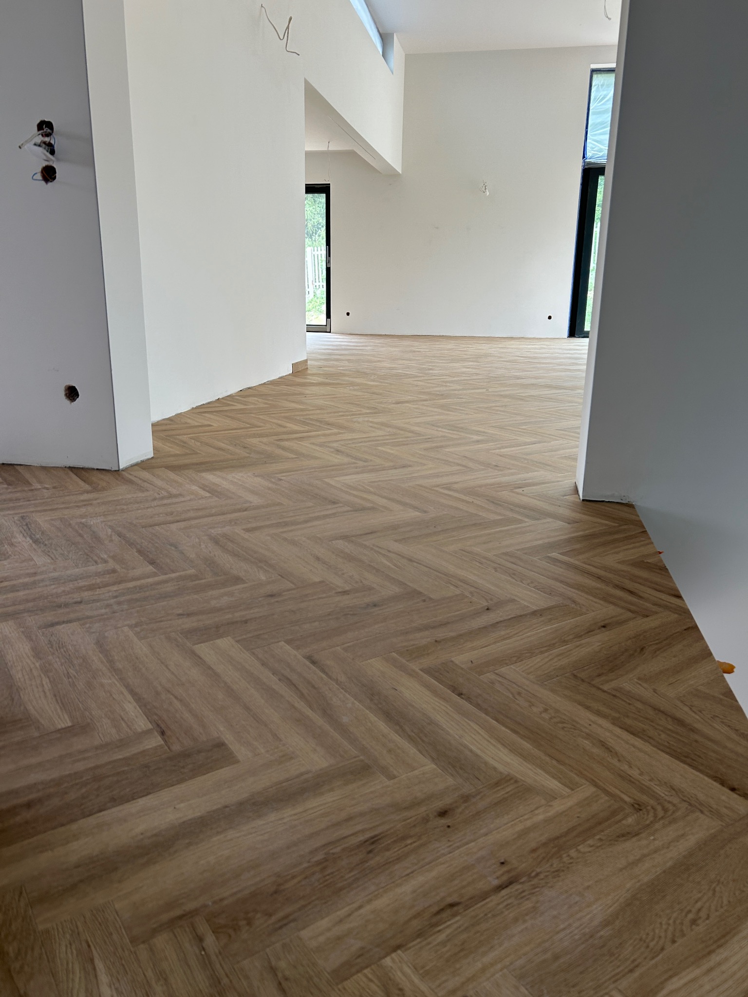 Ułożona jodełka z paneli podłogowych w surowym wnętrzu z białymi ścianami i widocznymi instalacjami elektrycznymi.