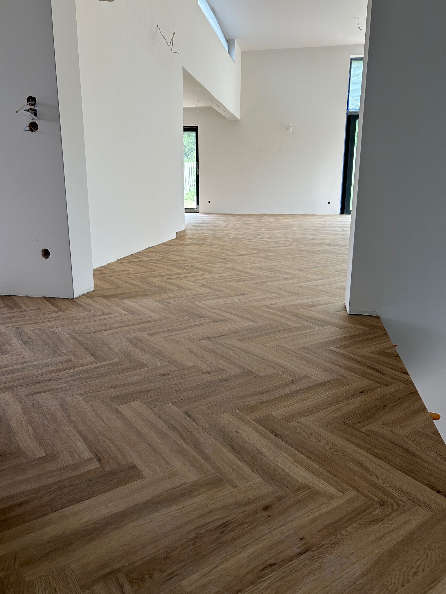 Jasnobrązowa podłoga ułożona we wzór jodełki w przestronnym, jasnym pomieszczeniu w trakcie wykańczania, z widocznymi otworami na gniazdka elektryczne i niezabezpieczonymi oknami.