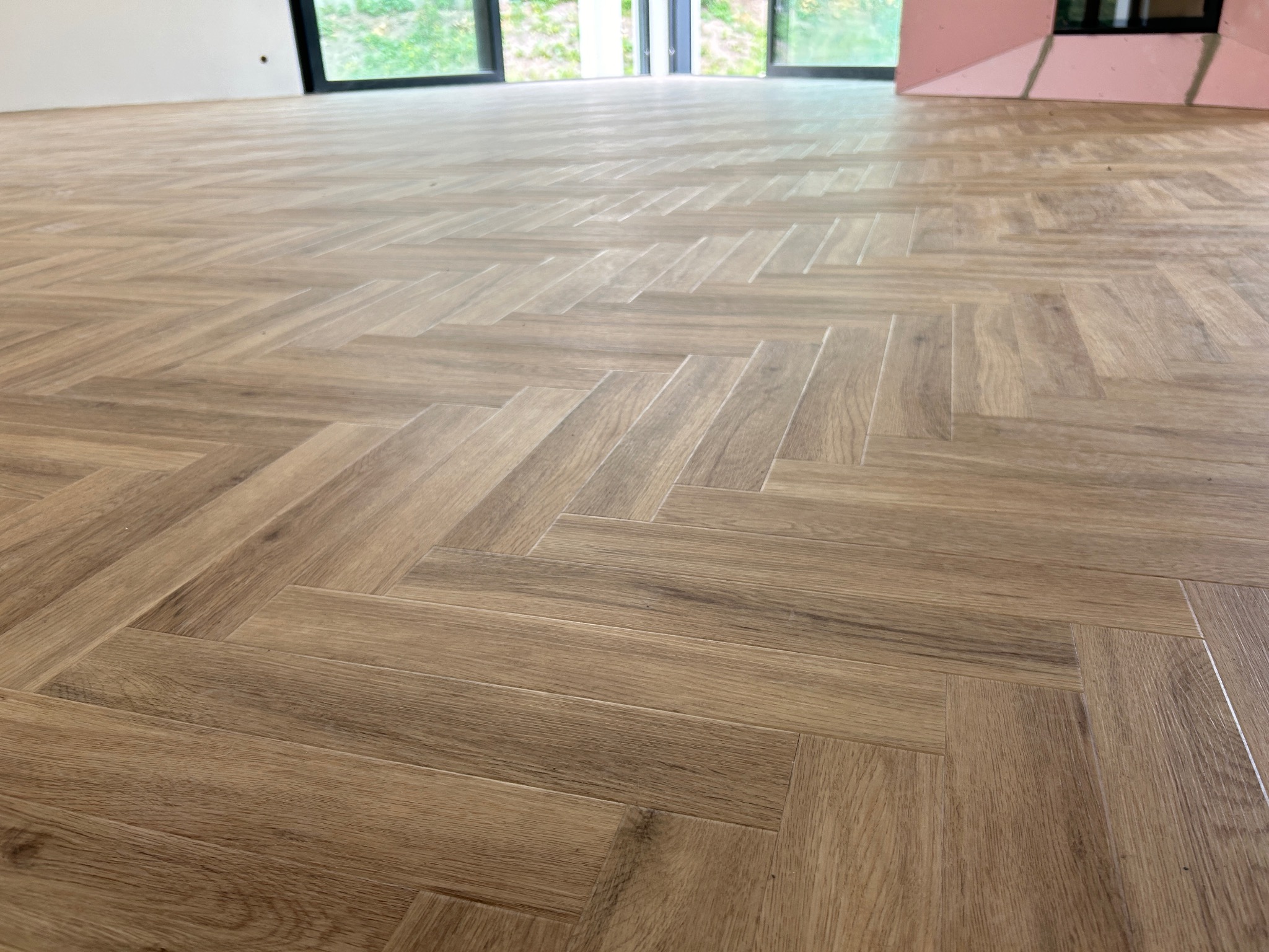 Ułożona wzorem jodełki, drewnopodobna podłoga w jasnym odcieniu brązu w nowym pomieszczeniu z dużymi oknami i fragmentem ściany w kolorze różowym w tle.