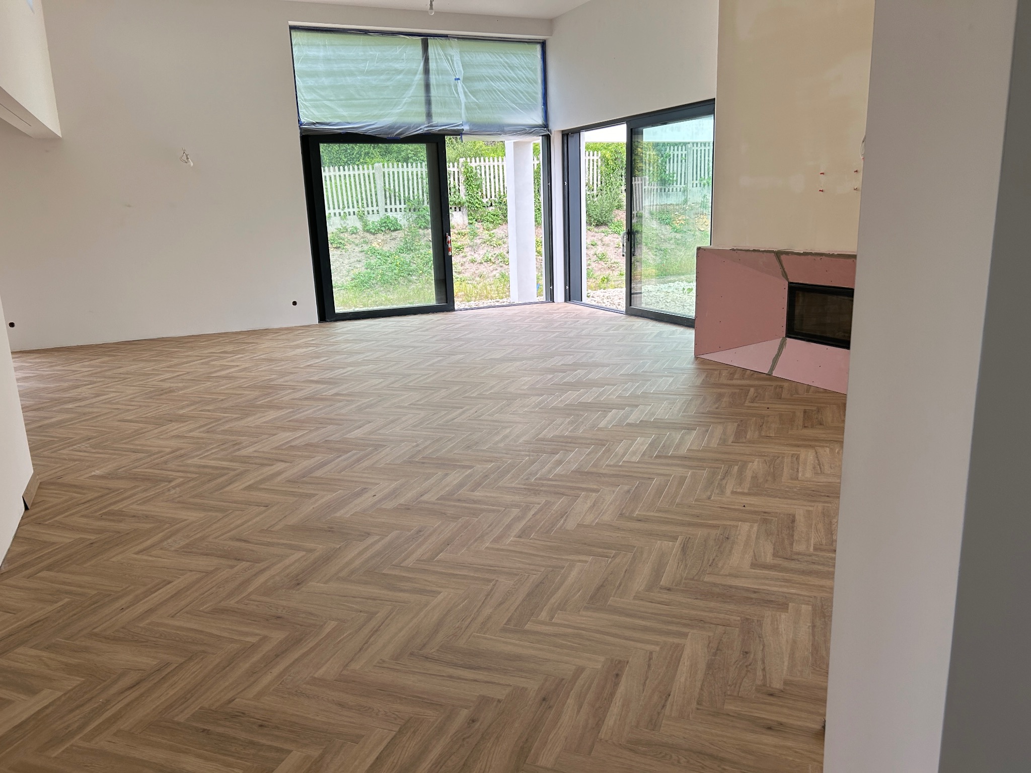 Nowoczesne wnętrze z ułożoną podłogą w jodełkę z paneli. Widoczne duże okna z widokiem na zieleń i częściowo zakryte folią. Kominek w trakcie wykończenia z płyt gipsowych.