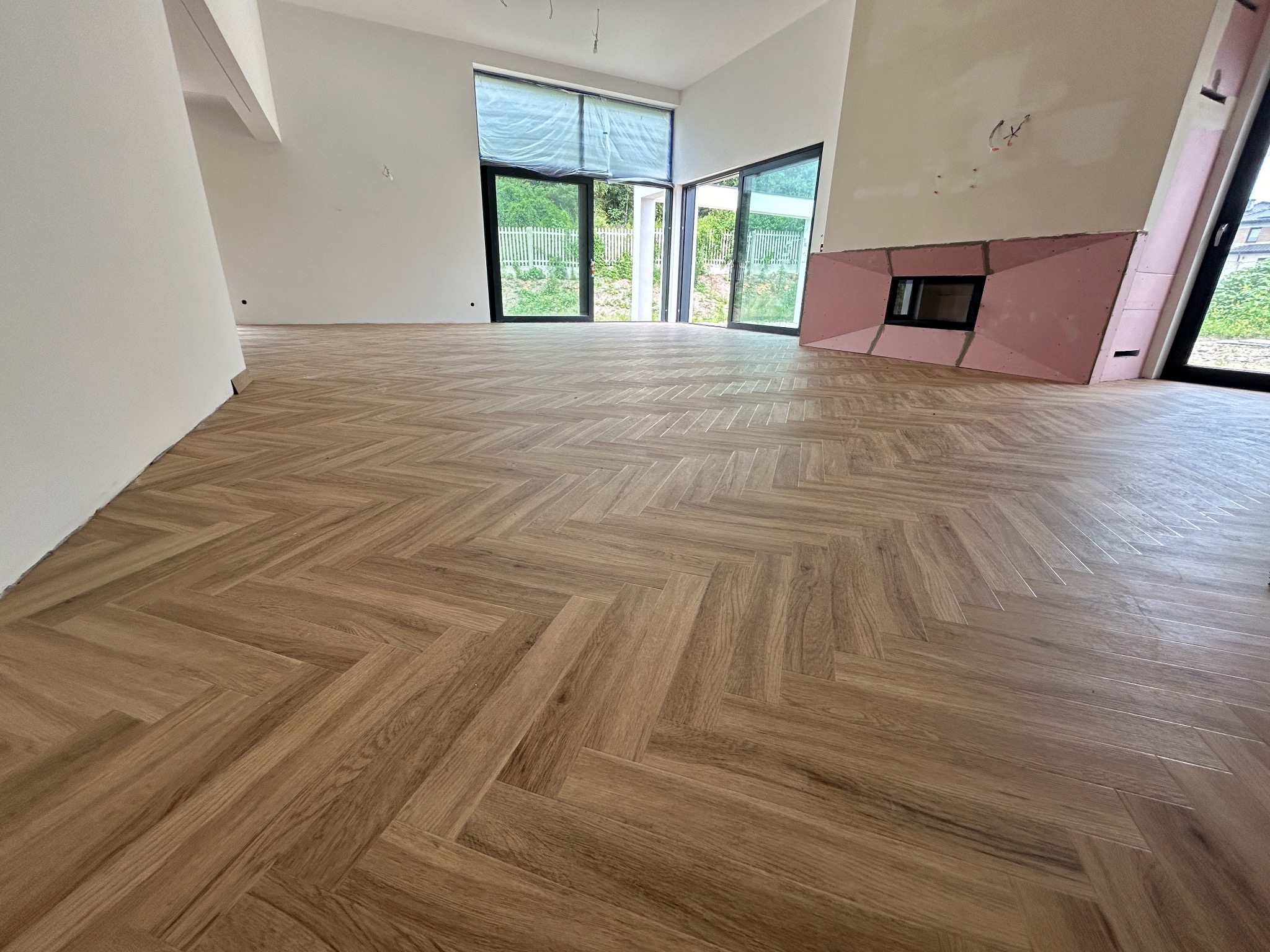 Jasna, drewniana podłoga ułożona w jodełkę w przestronnym, jasnym wnętrzu z dużymi oknami i przygotowanym miejscem na kominek.