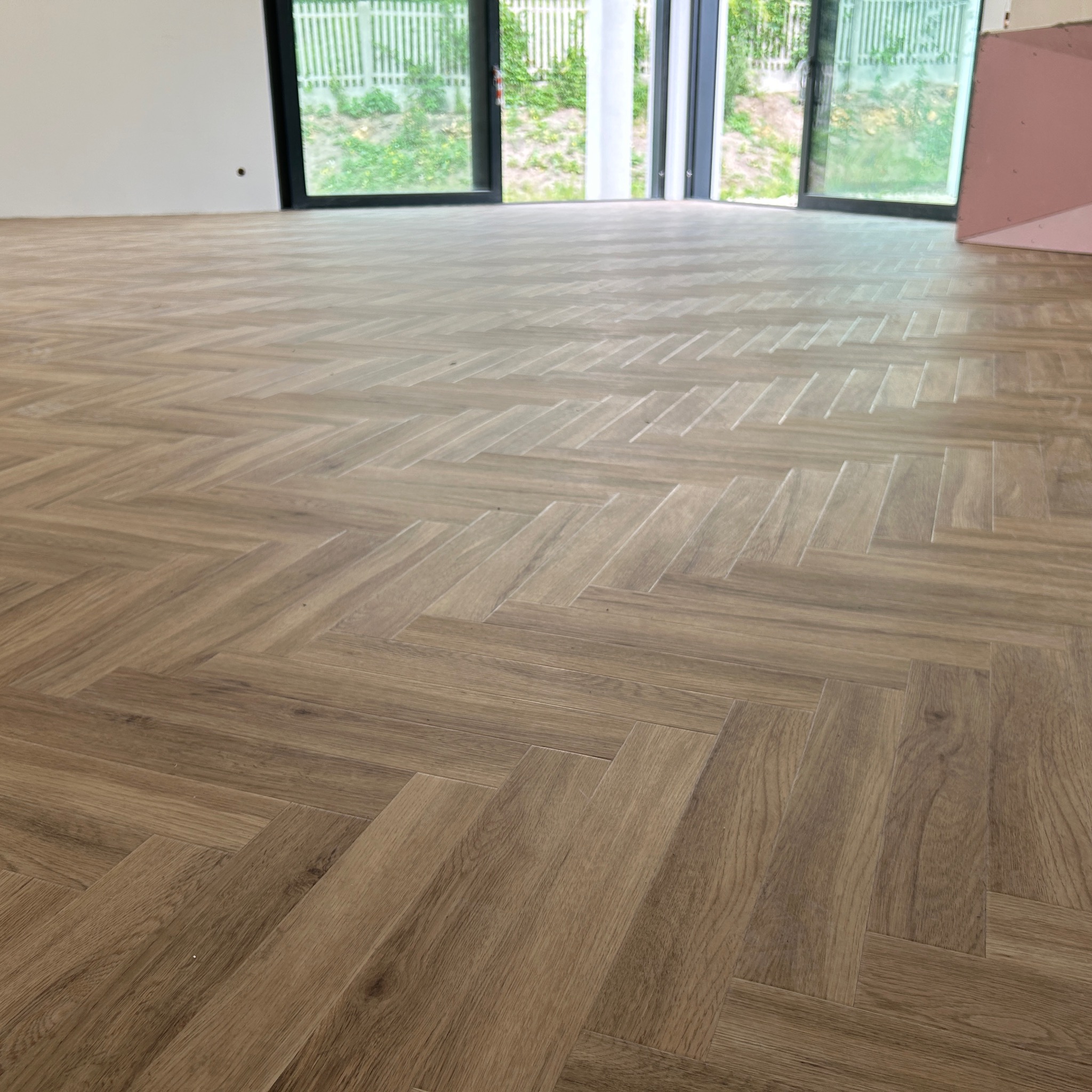 Ułożona podłoga z paneli w jodełkę w jasnym odcieniu drewna, widok na rozsuwane drzwi balkonowe i fragment ściany z płyt kartonowo-gipsowych.