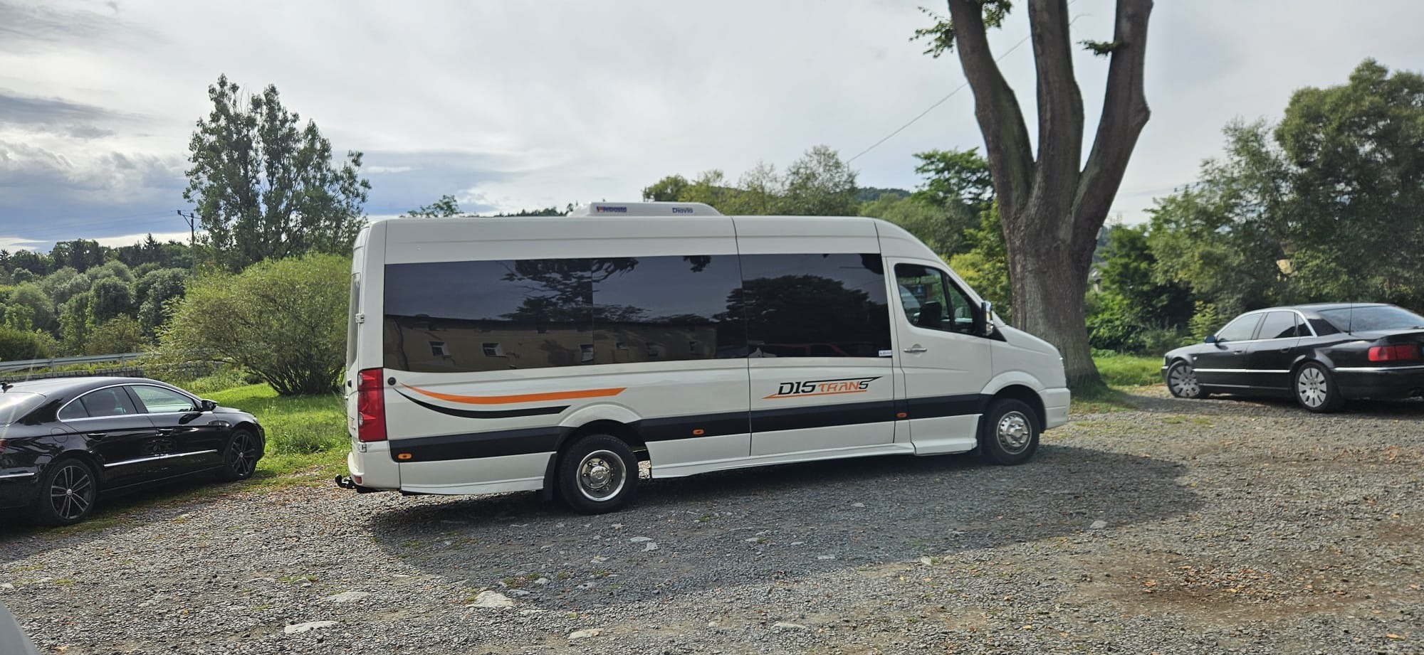 Biały bus z ciemnymi szybami i logo firmy DIS TRANS na tle zieleni i dwóch samochodów osobowych. Bus zaparkowany na żwirowym podłożu.