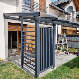 Rufin Beger - Szara, drewniana konstrukcja z zadaszeniem, prawdopodobnie wiata, na kostce brukowej, z drabiną w tle i zabawkami dziecięcymi na trawie.