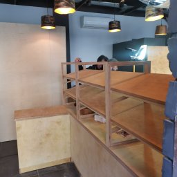 Rufin Beger - Montaż regału z jasnej sklejki w pomieszczeniu z oświetleniem punktowym; widoczna osoba mocująca elementy, ściana wyłożona płytami ze sklejki.