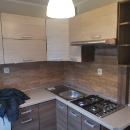 Rufin Beger - Komplet szafek kuchennych w jasnych odcieniach drewna z brązowym blatem, widoczna płyta gazowa i zlew, na blacie leży kurtka.