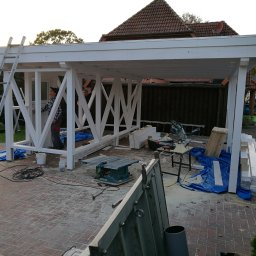 Rufin Beger - Budowa białej, drewnianej altany ogrodowej w trakcie realizacji, widoczne narzędzia, drabina i pracownicy w tle.