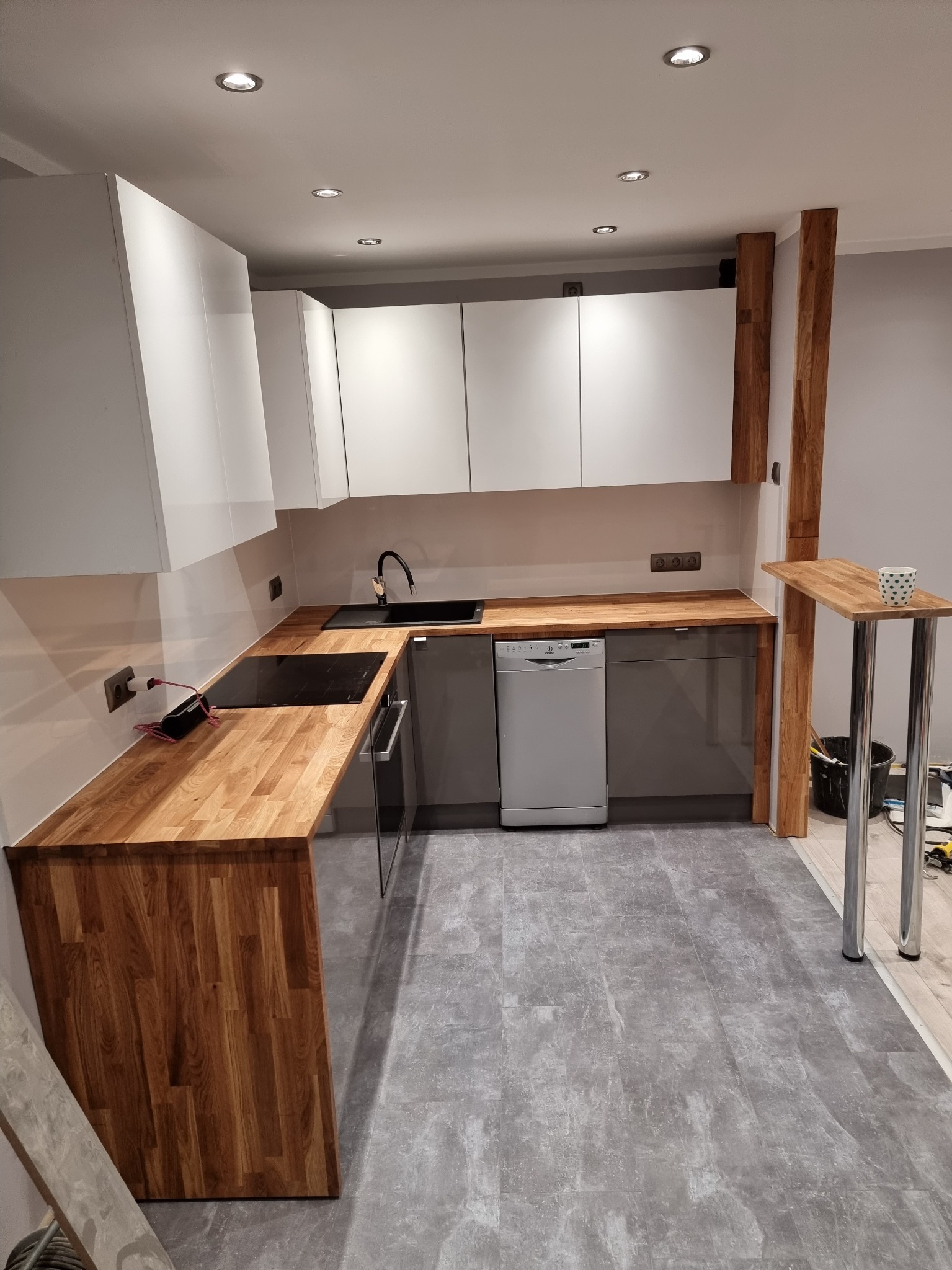 Nowoczesna kuchnia z białymi szafkami, drewnianym blatem i szarymi panelami podłogowymi, widoczny fragment baru z kubkiem w kropki.