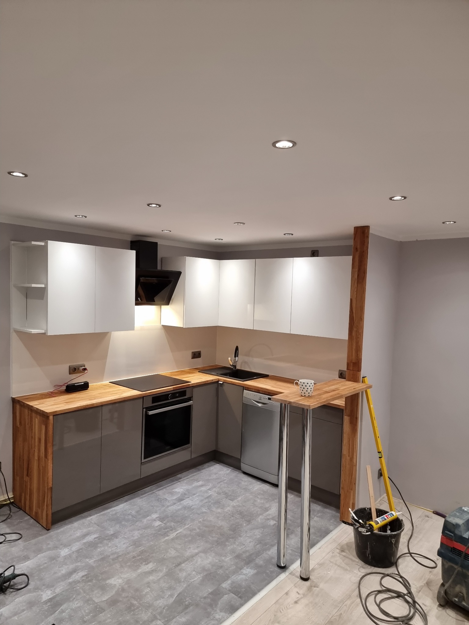 Nowoczesna kuchnia z szarymi i białymi szafkami, drewnianymi blatami, czarnym okapem, płytą indukcyjną, srebrną zmywarką i barkiem z drewnianym blatem na metalowych nogach, widoczne narzędzia...