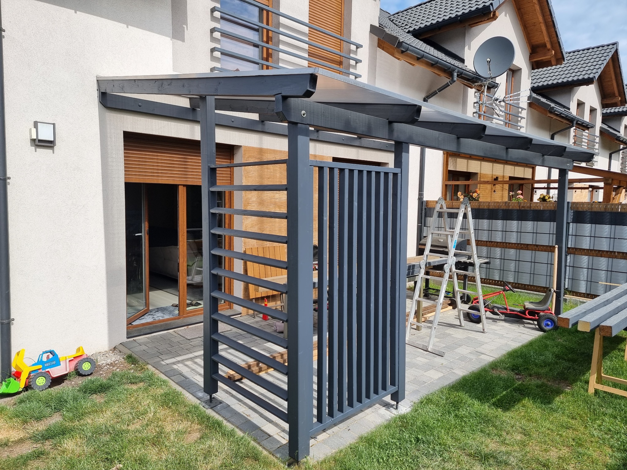 Szara, drewniana konstrukcja z zadaszeniem, prawdopodobnie wiata, na kostce brukowej, z drabiną w tle i zabawkami dziecięcymi na trawie.