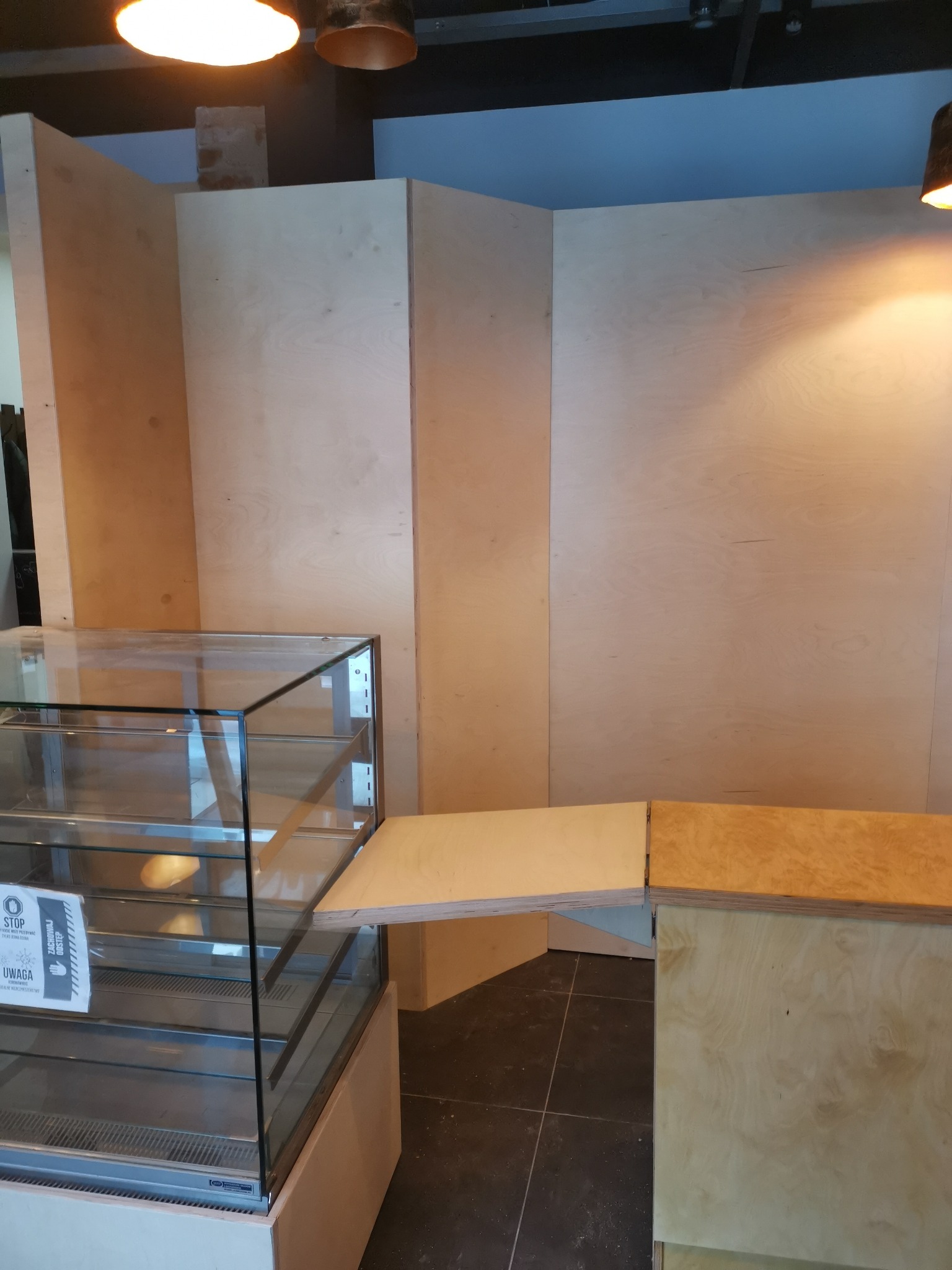 Montaż zabudowy z płyty drewnopodobnej w lokalu usługowym, widoczna przeszklona witryna chłodnicza i fragment sufitu z oświetleniem.