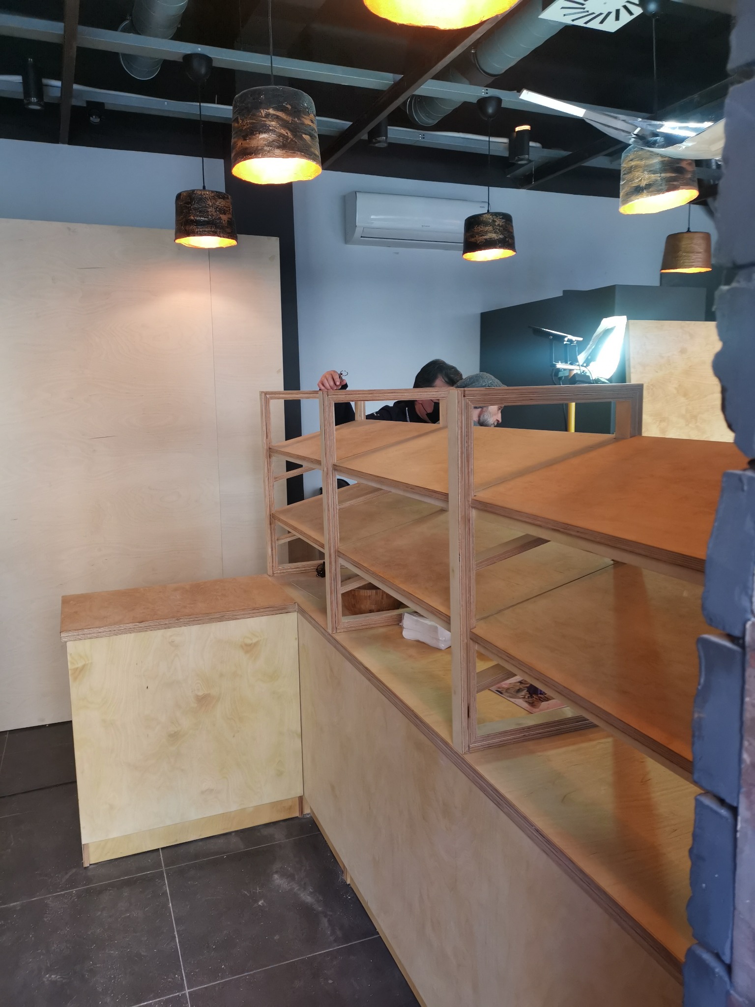 Montaż regału z jasnej sklejki w pomieszczeniu z oświetleniem punktowym; widoczna osoba mocująca elementy, ściana wyłożona płytami ze sklejki.