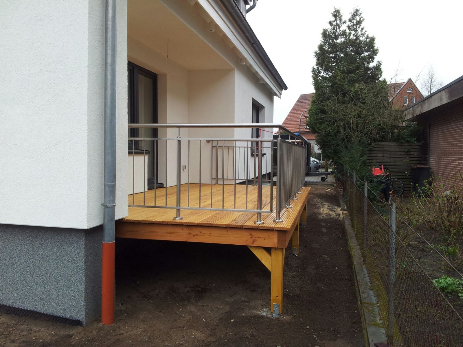 Nowoczesny taras z drewnianą podłogą i metalową balustradą, widok z zewnątrz na tle białego budynku z szarą elewacją przyziemia i fragmentu ogrodu.