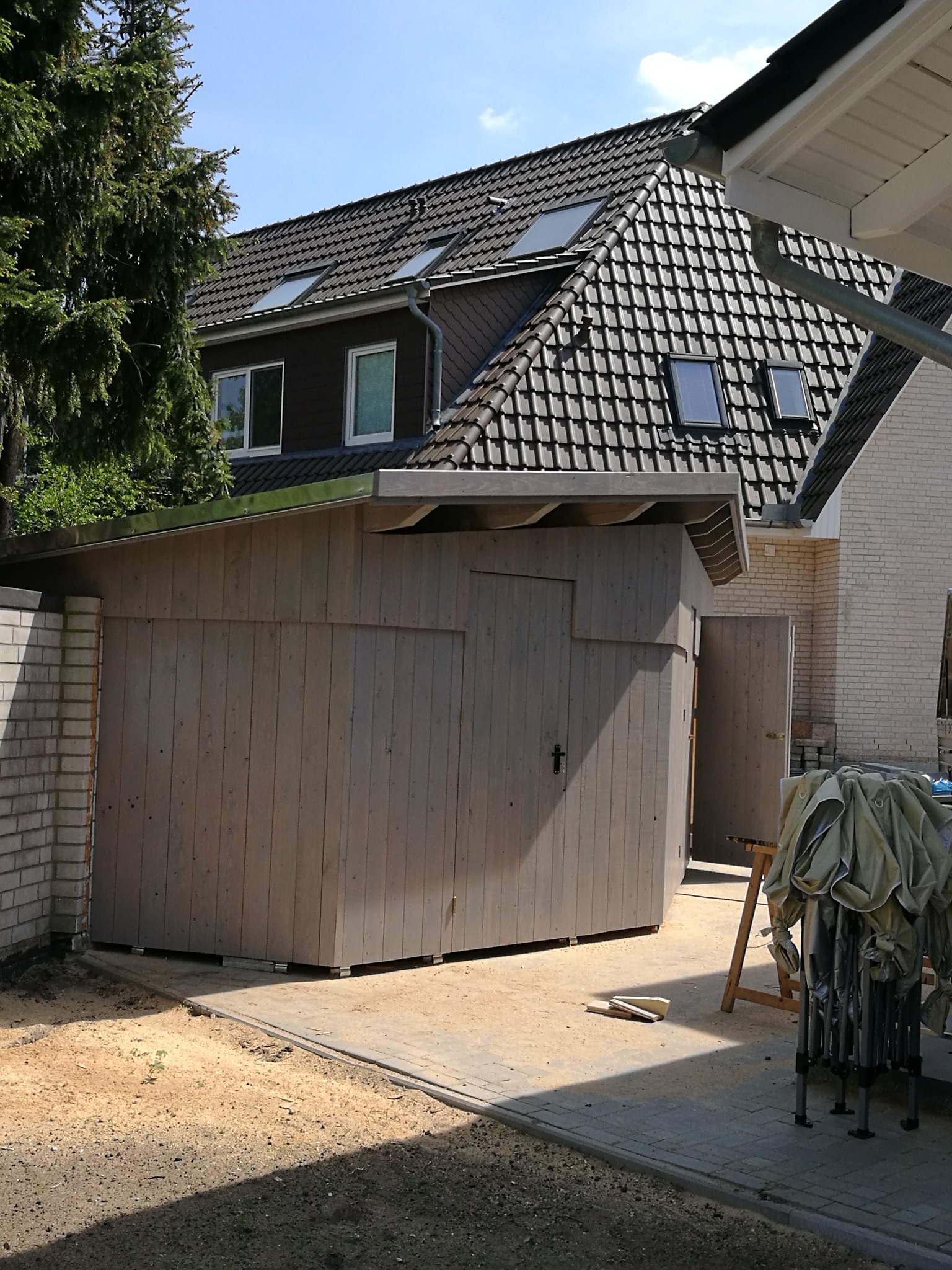 Szara, drewniana konstrukcja garażowa z otwartymi drzwiami, przylegająca do murowanego budynku, na tle domu z ciemną dachówką i oknami dachowymi.
