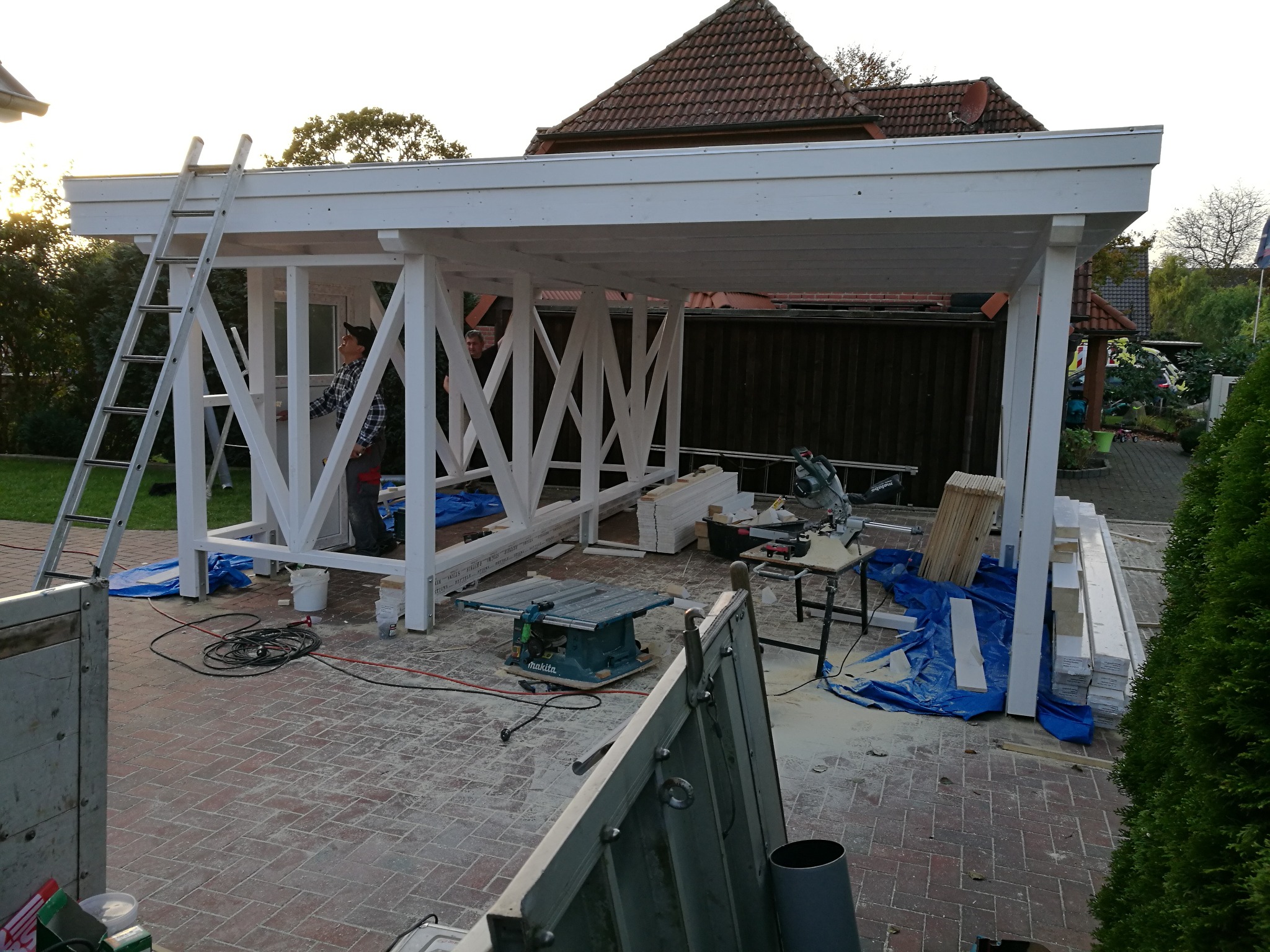Budowa białej, drewnianej altany ogrodowej w trakcie realizacji, widoczne narzędzia, drabina i pracownicy w tle.