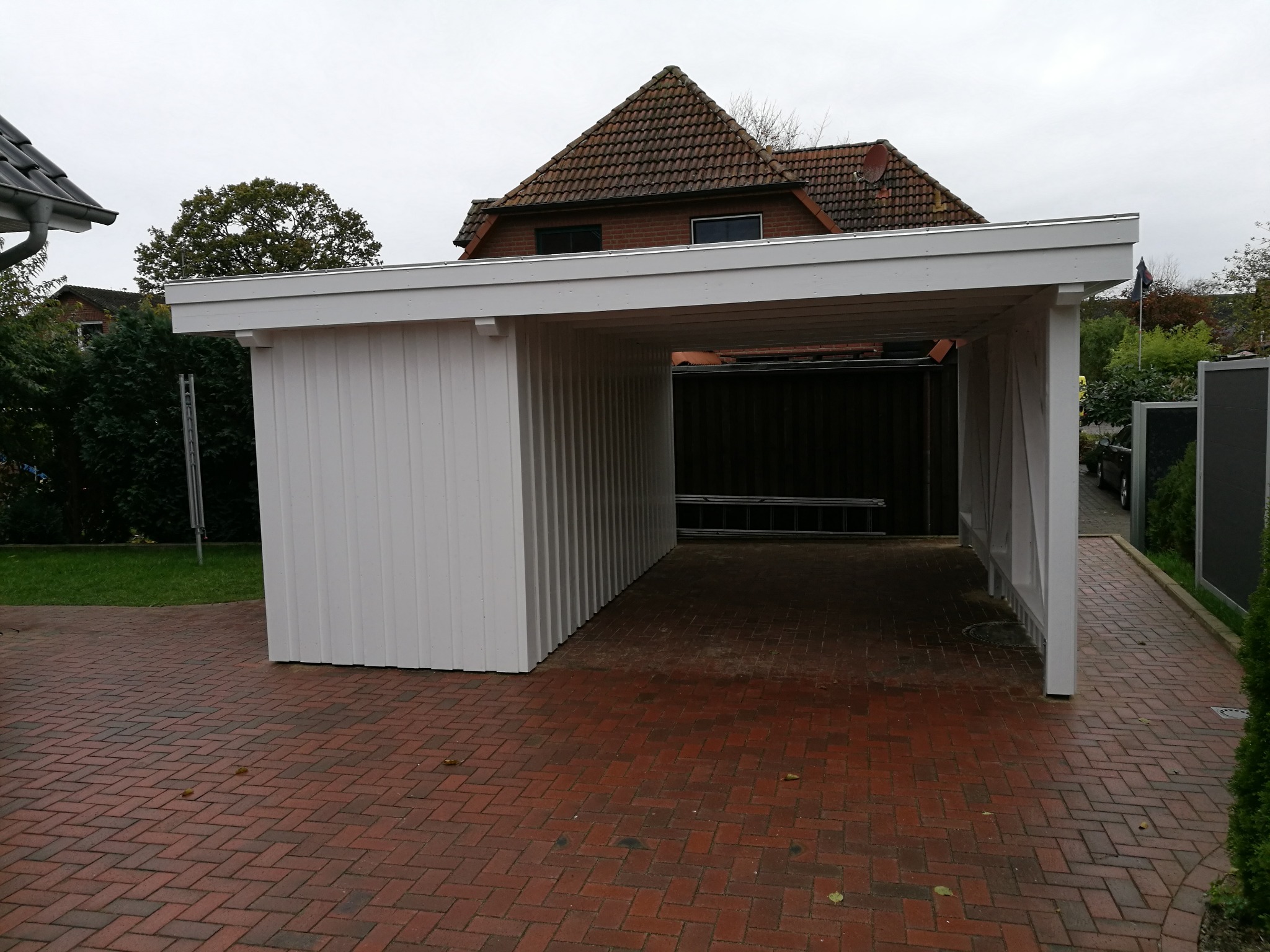 Nowo wybudowana, biała, drewniana wiata garażowa z widoczną konstrukcją dachu i ceglaną nawierzchnią podjazdu, na tle domu z czerwonej cegły i zieleni.