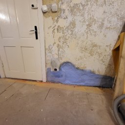 KB EFEKT Bartek Kartuszyński - Przygotowanie ściany do prac wykończeniowych: fragment ściany z odpryskującą farbą, zagruntowany na niebiesko dolny pas, widoczne ślady po instalacji elektrycznej, obok białe drzwi i fragment...