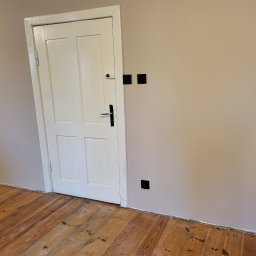 KB EFEKT Bartek Kartuszyński - Świeżo pomalowane wnętrze z drewnianą podłogą, białą ścianą z czarnymi gniazdkami i fragmentem ceglanego kominka.