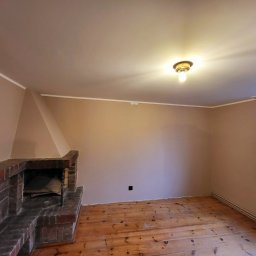 KB EFEKT Bartek Kartuszyński - Pokój po odświeżeniu ścian w odcieniach beżu, z ceglanym kominkiem, drewnianą podłogą i grzejnikiem pod oknem.