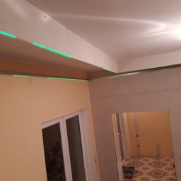 KB EFEKT Bartek Kartuszyński - Montaż płyt kartonowo-gipsowych na ścianie i suficie, widoczne zielone linie lasera wyznaczające poziom, otwór drzwiowy w tle.