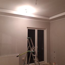 KB EFEKT Bartek Kartuszyński - Pokój po szpachlowaniu ścian i sufitu z widoczną białą sztukaterią sufitową, grzejnikiem i drabiną malarską z wałkiem.