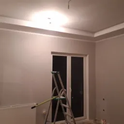 Pok&oacute;j po szpachlowaniu ścian i sufitu z widoczną białą sztukaterią sufitową, grzejnikiem i drabiną malarską z wałkiem.