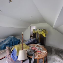 KB EFEKT Bartek Kartuszyński - Poddasze po malowaniu na biało, widoczne kable oświetleniowe, meble pod folią i stół z narzędziami. Ściany i sufit świeżo pomalowane, brak lamp.