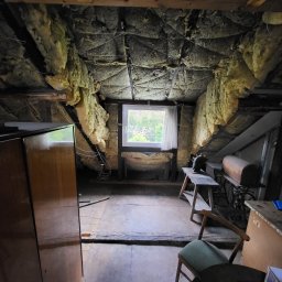KB EFEKT Bartek Kartuszyński - Poddasze z oknem i izolacją ścian wełną mineralną, widoczne stare meble i elementy konstrukcyjne dachu. Ujęcie z perspektywy podłogi, z widocznym oknem w tle.