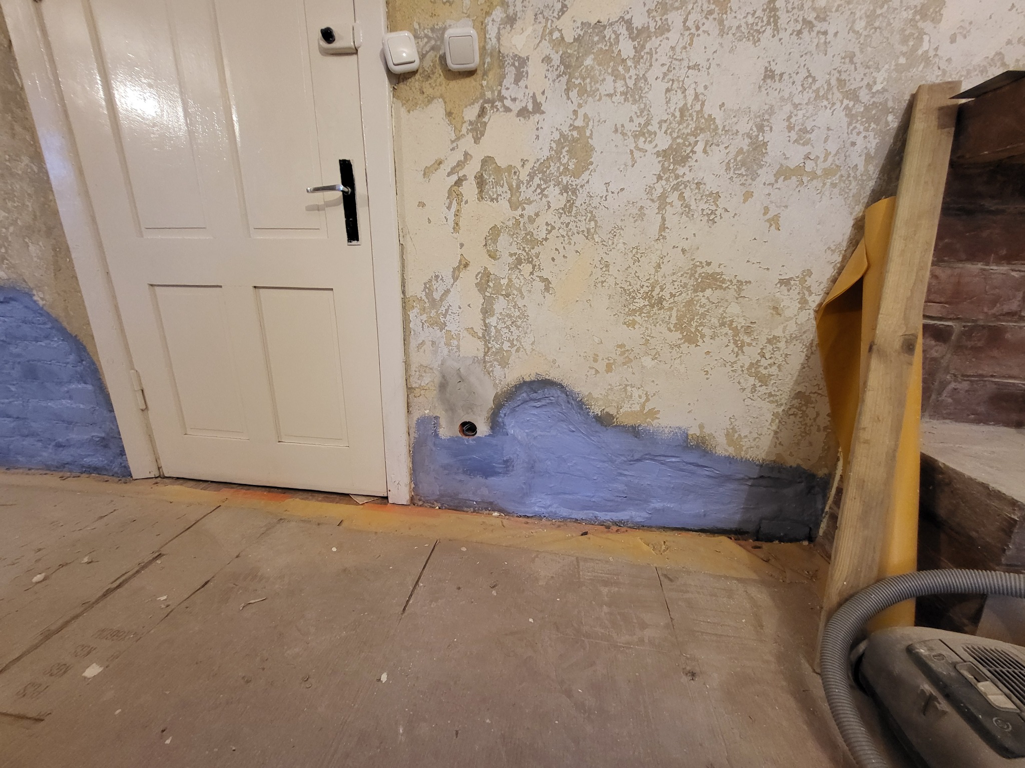 Przygotowanie ściany do prac wykończeniowych: fragment ściany z odpryskującą farbą, zagruntowany na niebiesko dolny pas, widoczne ślady po instalacji elektrycznej, obok białe drzwi i fragment...