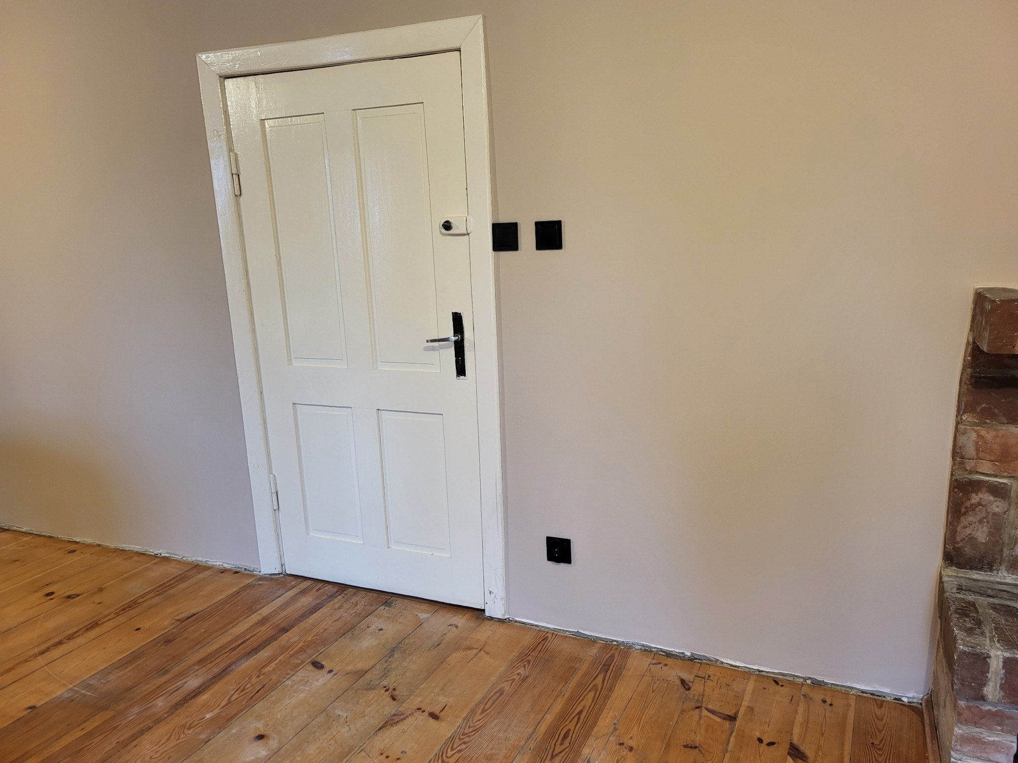 Świeżo pomalowane wnętrze z drewnianą podłogą, białą ścianą z czarnymi gniazdkami i fragmentem ceglanego kominka.