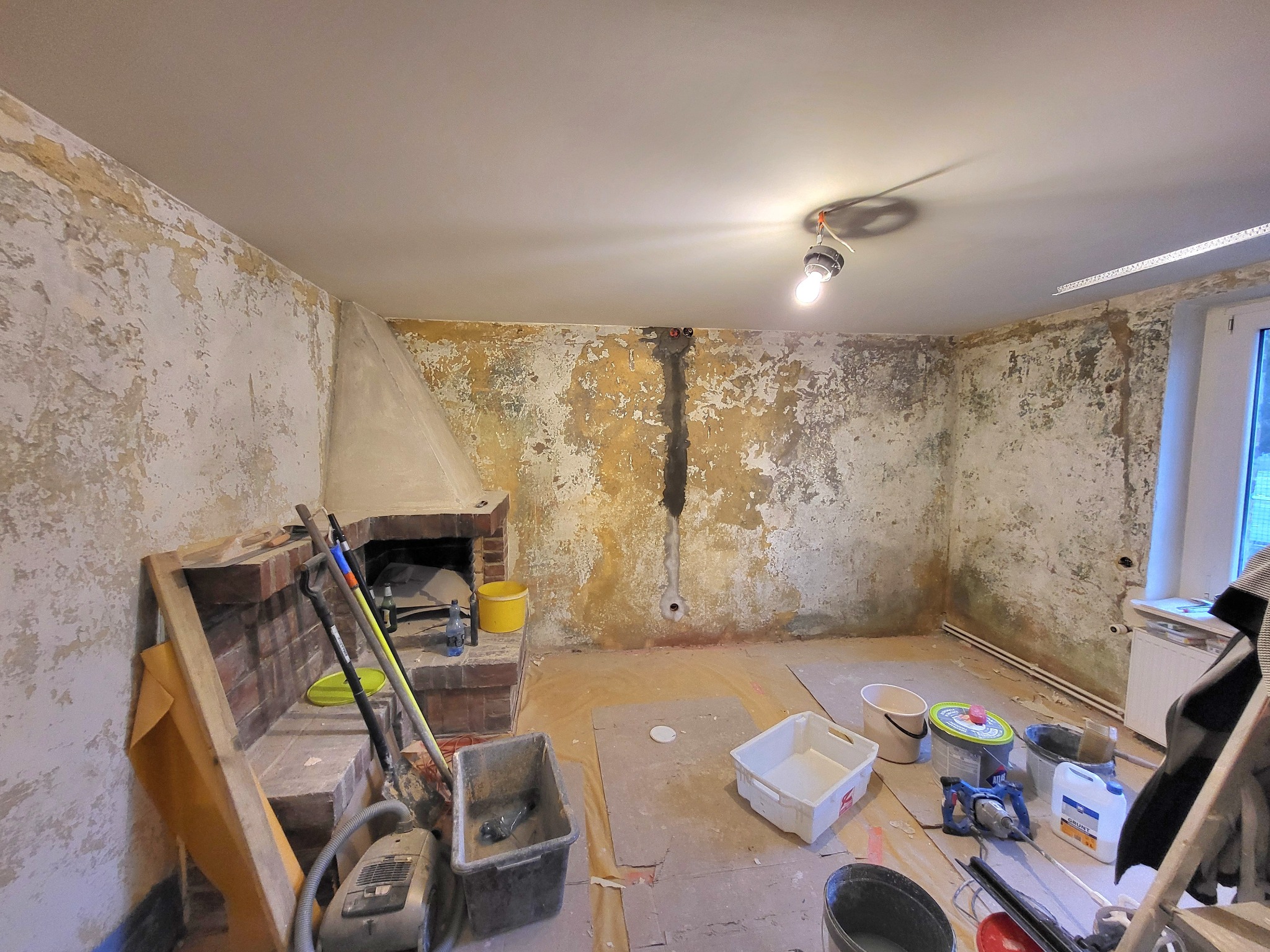 Pokój w trakcie remontu: ściany ze śladami starej farby i tapet, widoczny kominek, wiaderka, narzędzia, podłoga zabezpieczona kartonem, surowa żarówka.