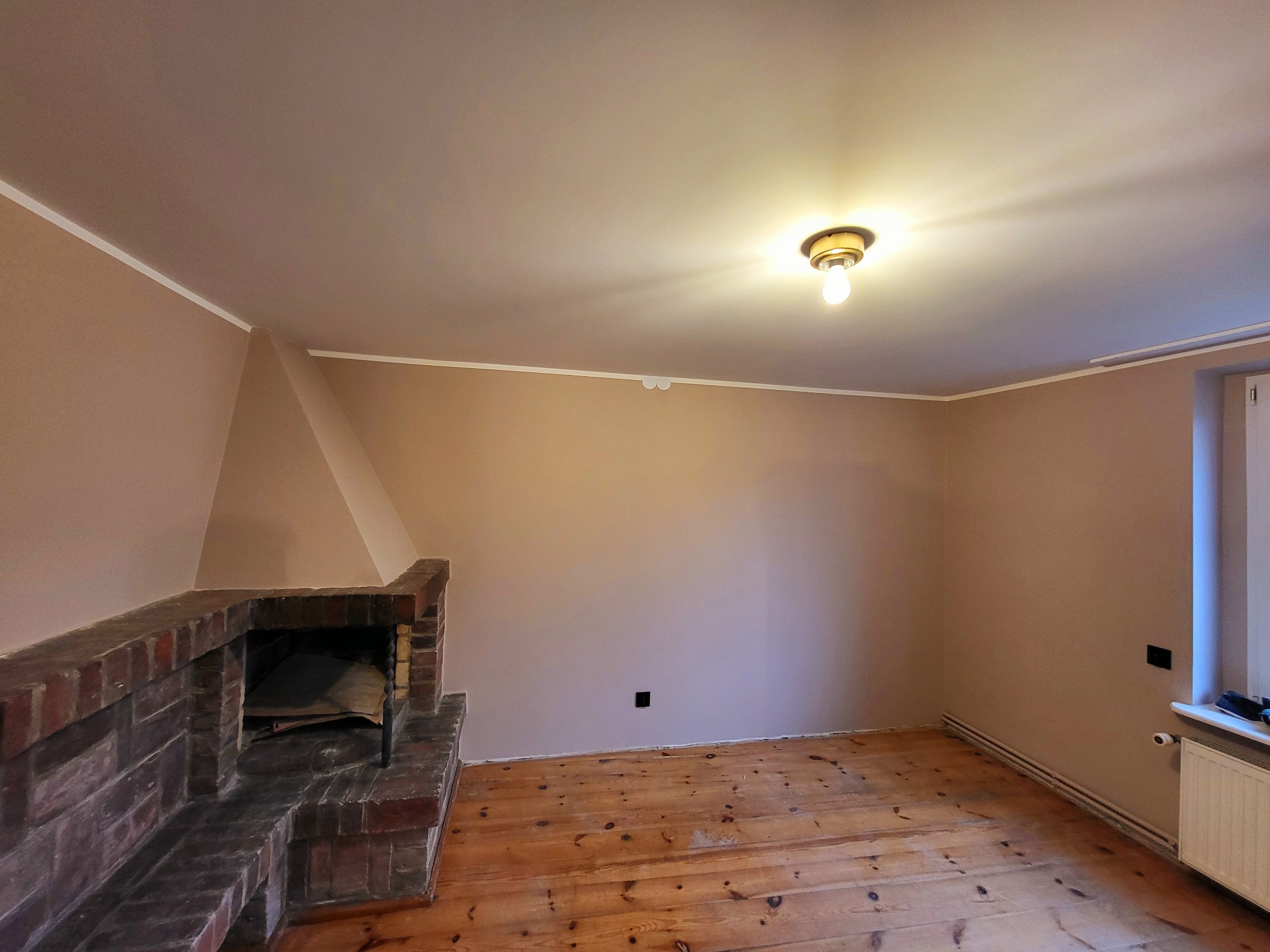 Pokój po odświeżeniu ścian w odcieniach beżu, z ceglanym kominkiem, drewnianą podłogą i grzejnikiem pod oknem.