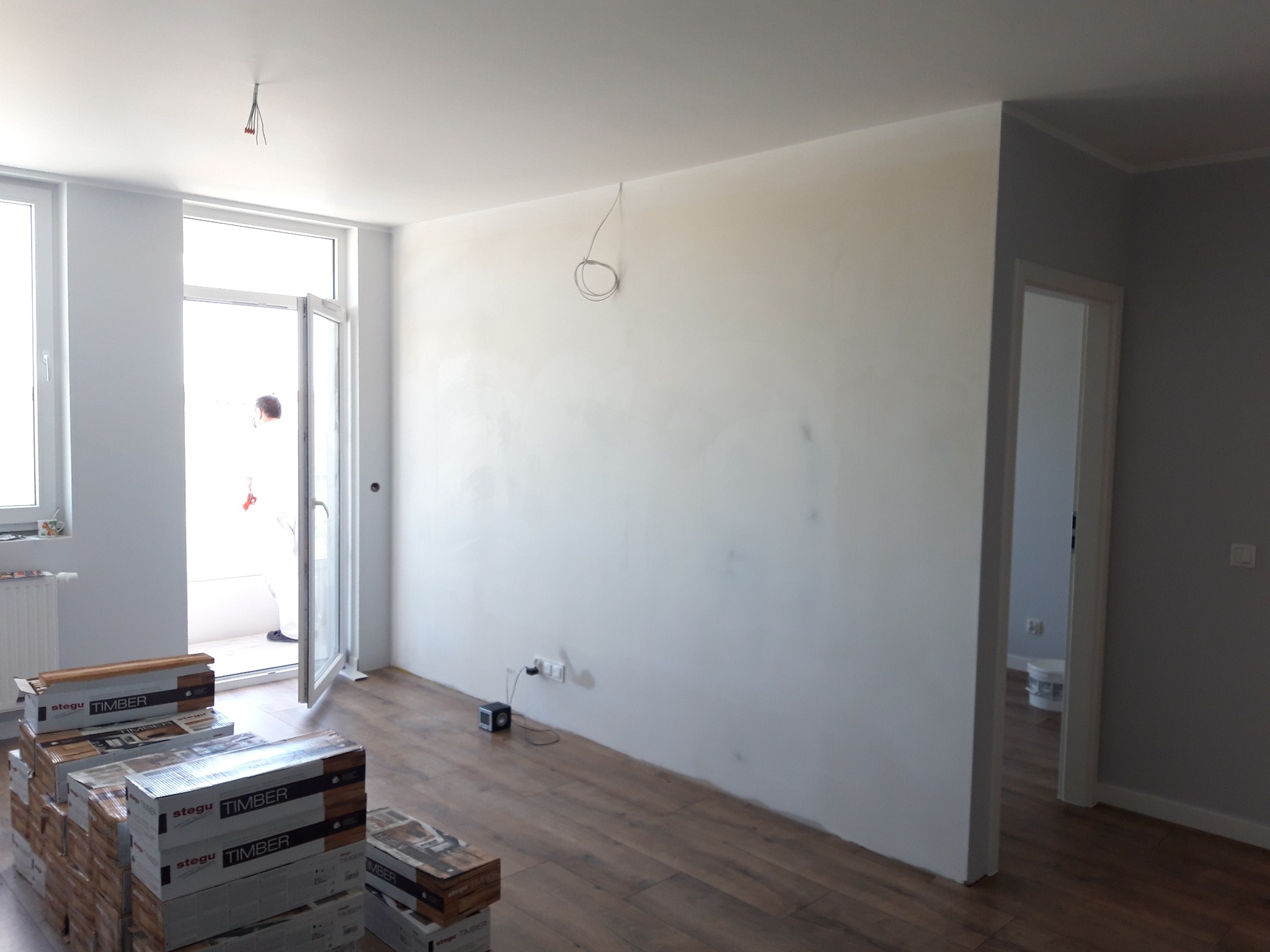 Pokój w trakcie wykańczania: świeżo położone panele podłogowe, ściany po wstępnym wyrównaniu, widoczne puszki paneli Stegu Timber, wystające przewody elektryczne z sufitu, otwarte drzwi balkonowe...