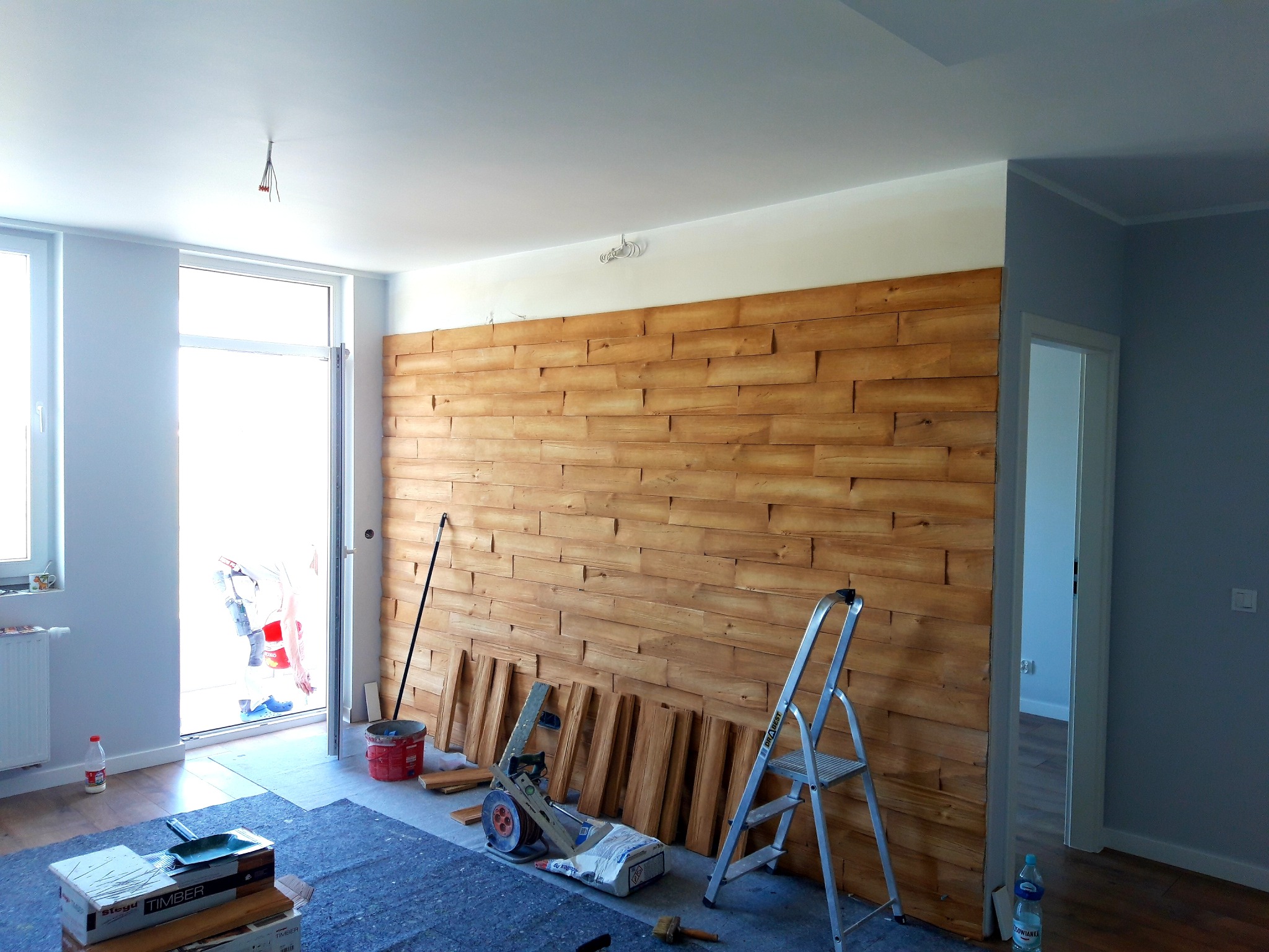 Montaż dekoracyjnej ściany z paneli drewnianych, widoczne narzędzia, drabina aluminiowa, poziomica i materiały budowlane, pomieszczenie w trakcie wykończenia.