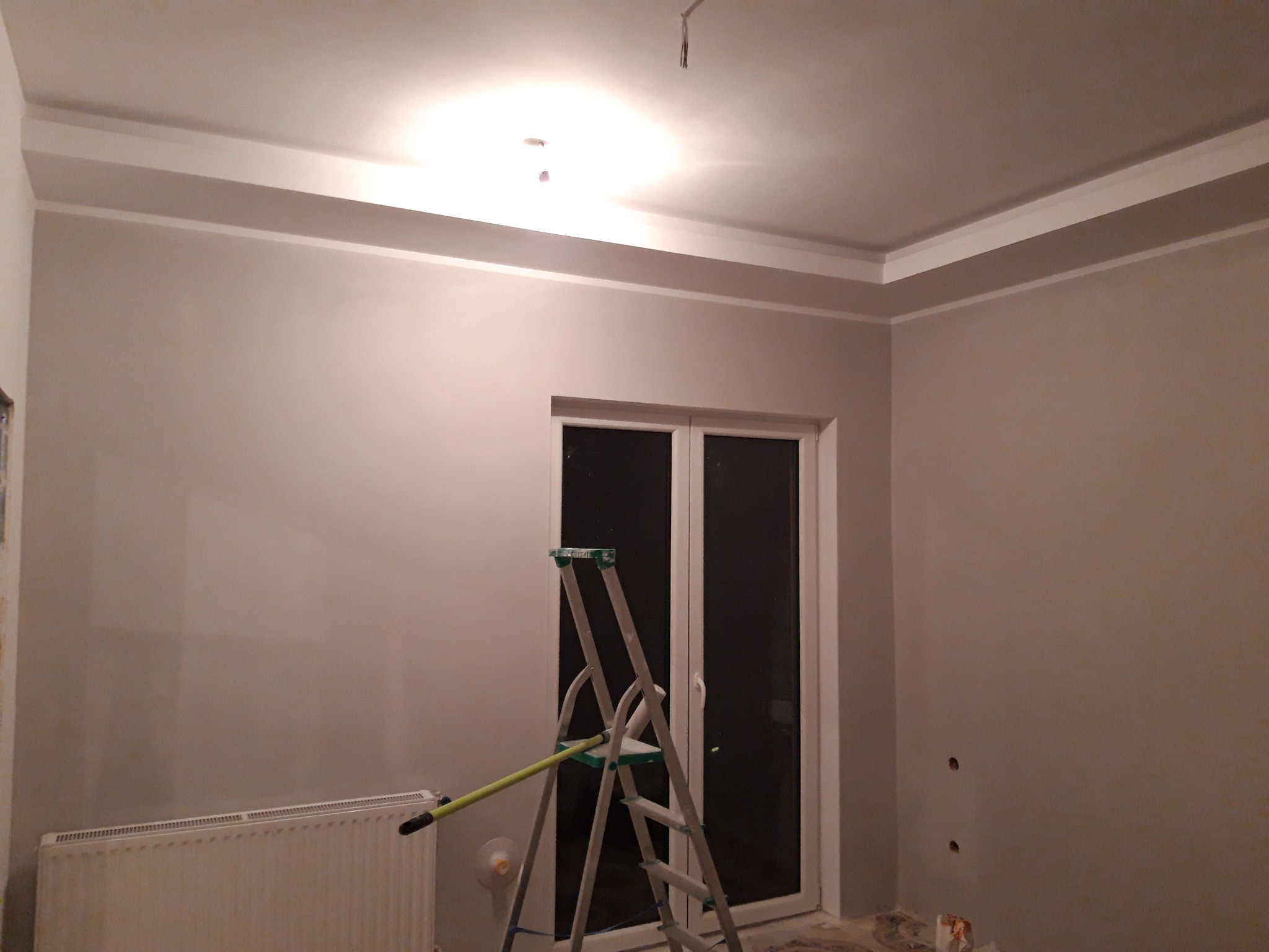 Pokój po szpachlowaniu ścian i sufitu z widoczną białą sztukaterią sufitową, grzejnikiem i drabiną malarską z wałkiem.
