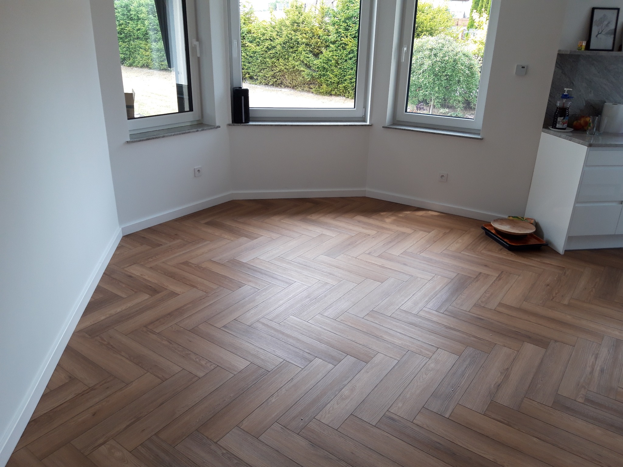 Ułożone panele podłogowe w jodełkę w jasnym pomieszczeniu w Dziemianach, widok na okna z zielenią na zewnątrz.