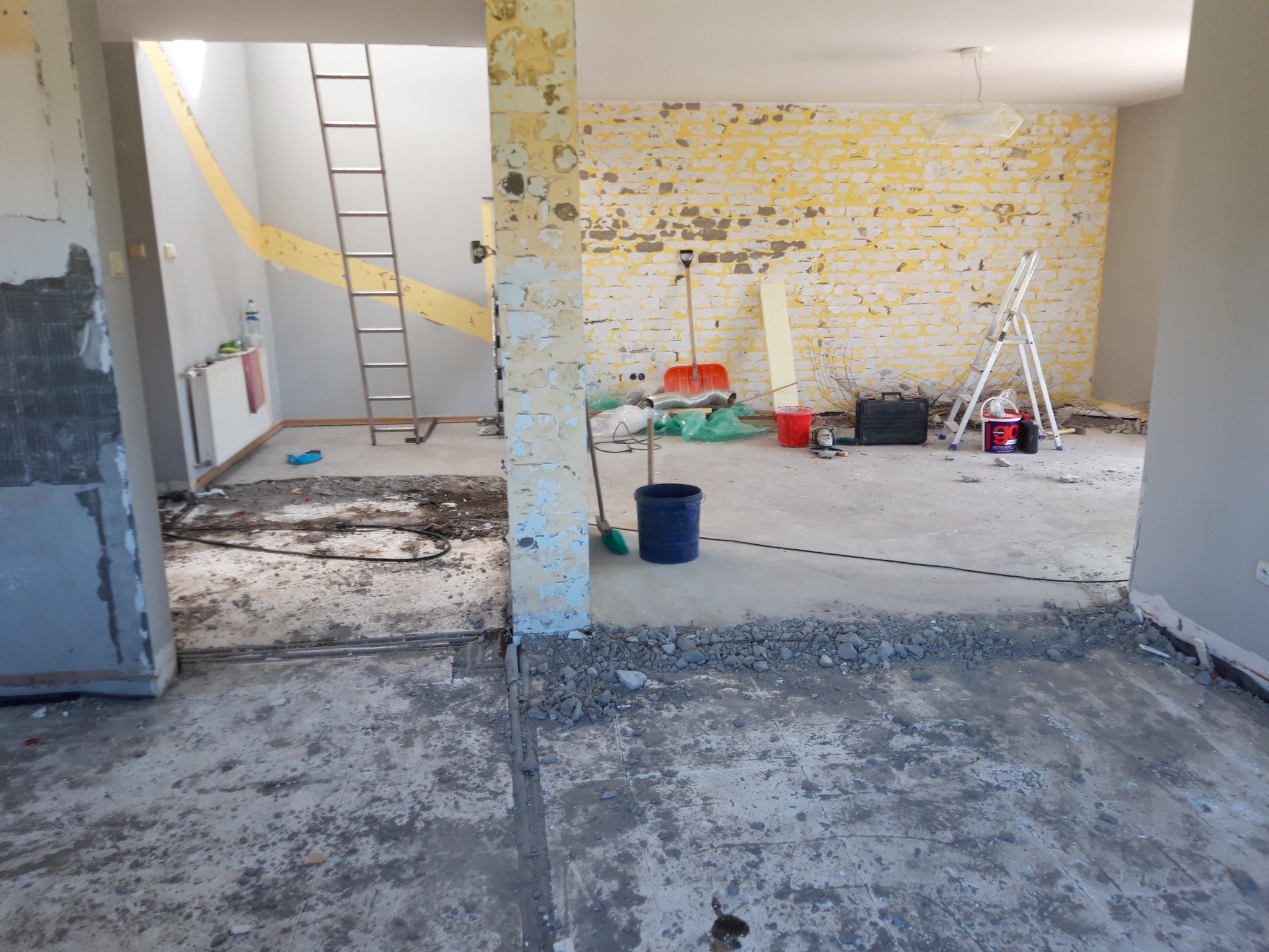 Wnętrze w trakcie prac remontowych: ściany z resztkami farby, gruz na podłodze, drabina oparta o ścianę, widoczne narzędzia i akcesoria malarskie.