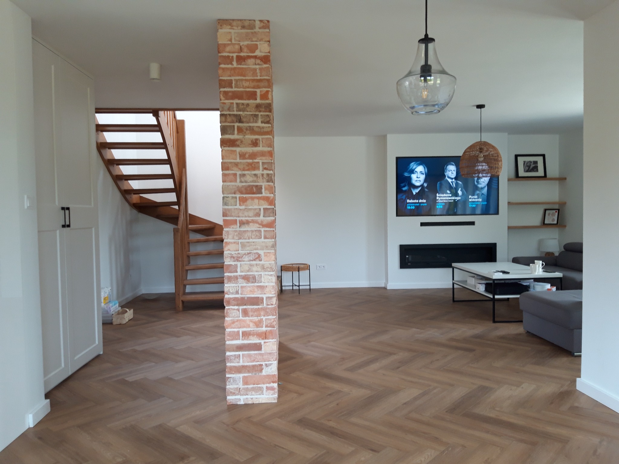 Salon po remoncie z podłogą ułożoną w jodełkę, ceglanym filarem i schodami drewnianymi, nowoczesny wystrój.