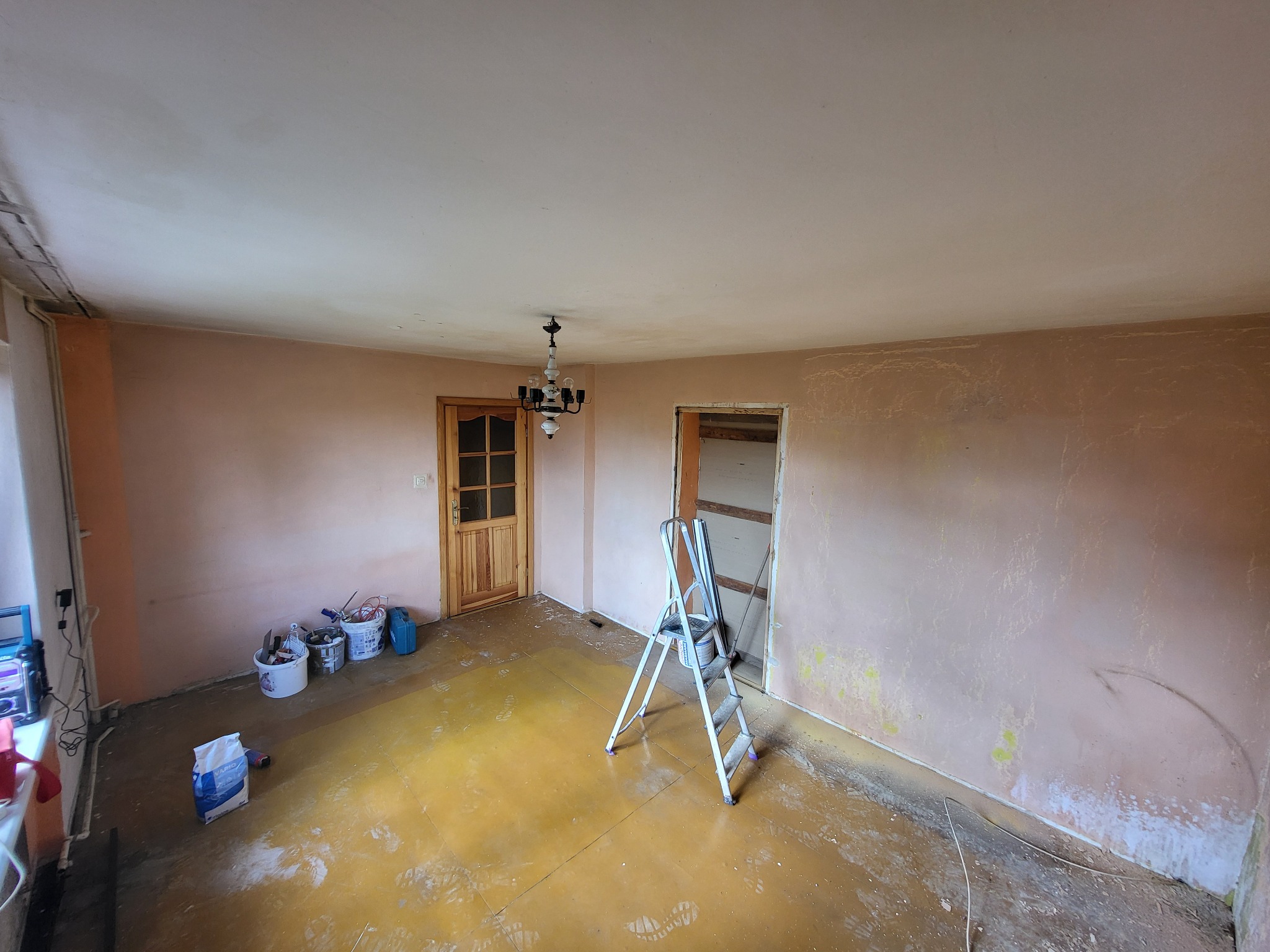 Pokój w trakcie remontu: ściany po szpachlowaniu, przygotowane do malowania, widoczne narzędzia malarskie, drabina aluminiowa i zabezpieczona folią podłoga.