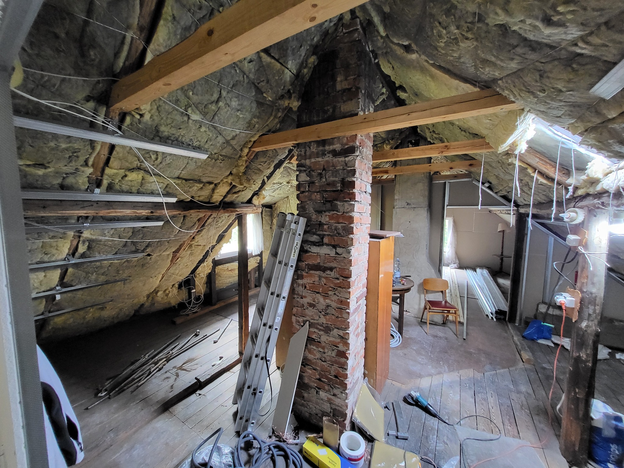 Poddasze w trakcie adaptacji: widoczne ocieplenie, konstrukcja drewniana, komin z cegły, drabina i narzędzia. Prace wykończeniowe w surowym stanie.