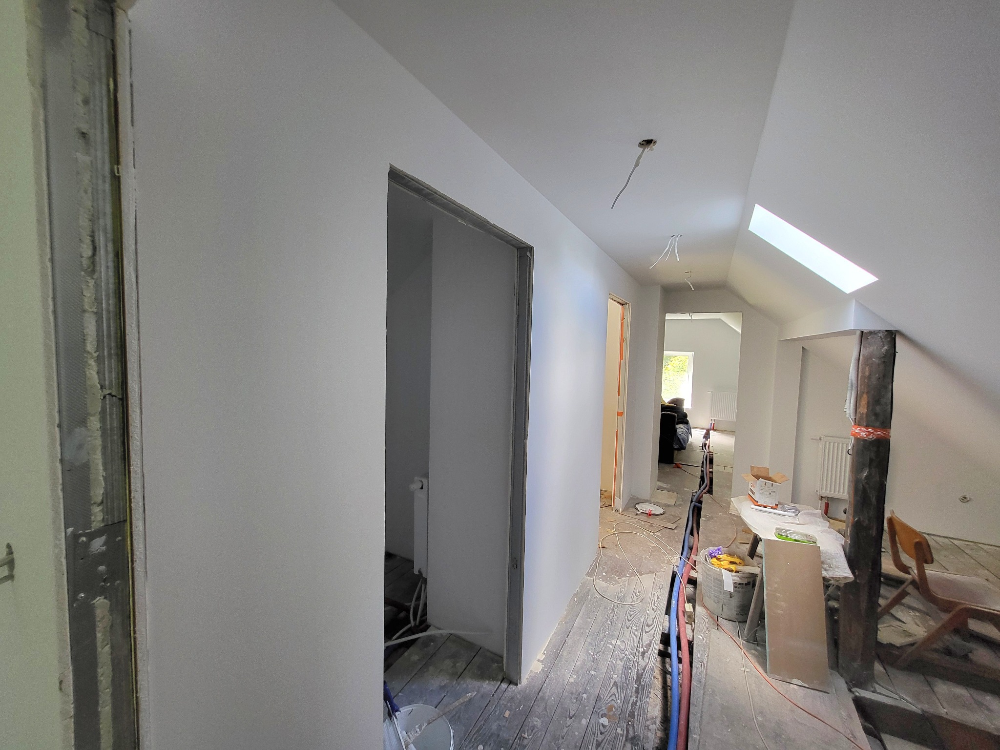 Białe, świeżo pomalowane ściany i sufit na poddaszu z oknem dachowym. Widoczne przygotowania do montażu drzwi i niedokończone podłogi.